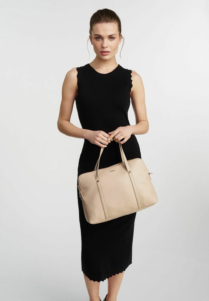 Beżowa skórzana torba na ramię z dwoma uchwytami, zamknięciem na zamek i pionowymi detalami szycia. Modelka ma na sobie czarną sukienkę bez rękawów z falbaniastym dołem.