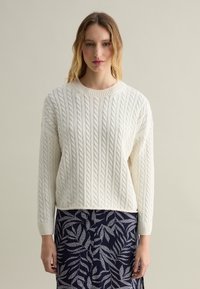 Cremfarbener Cable-Knit-Pullover mit Rundhalsausschnitt und langen �Ärmeln, kombiniert mit einem marineblauen Rock mit weißem Blattmuster.