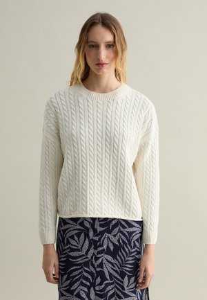 Suéter de punto trenzado color crema con cuello redondo y mangas largas, combinado con una falda azul marino con un patrón de hojas blancas.
