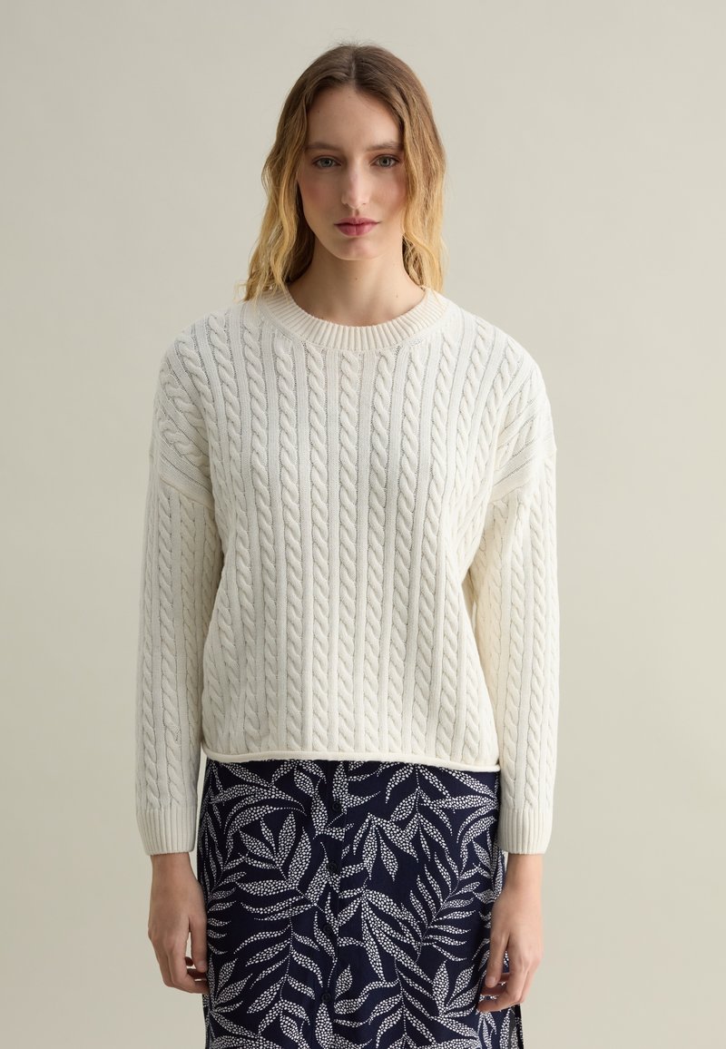 Cremfarbener Cable-Knit-Pullover mit Rundhalsausschnitt und langen Ärmeln, kombiniert mit einem marineblauen Rock mit weißem Blattmuster.