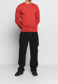 Sweat-shirt rouge à col rond avec manches longues ; associé à un pantalon noir avec coutures visibles et poches. Des chaussures de sport noires complètent la tenue.