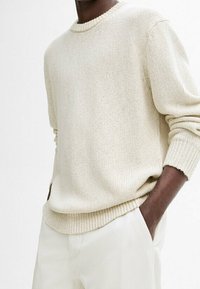 Maglione lavorato a maglia beige chiaro con scollatura rotonda, polsini e orlo a coste, abbinato a pantaloni chiari con vestibilità comoda.
