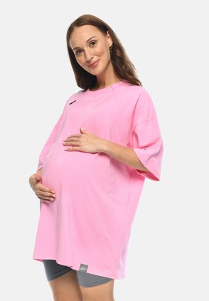 Schwangere Frau mit langen braunen Haaren, die ein lockeres rosa Shirt und graue Shorts trägt, hält sanft ihren Bauch und lächelt.