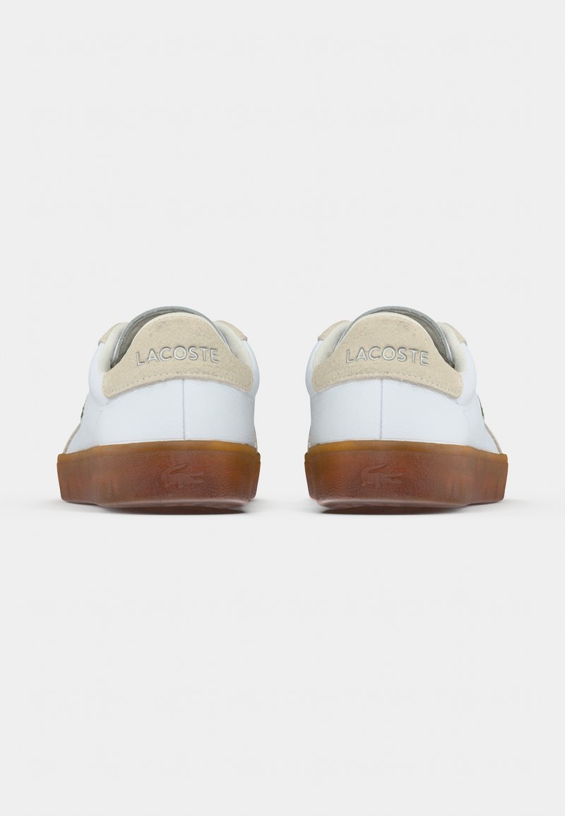 White sneakers with a textured beige suede heel, rubber sole, and "Lacoste" branding on the back. Minimalist design, smooth leather body.