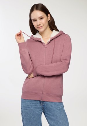 Felpa con zip rosa, foderata con morbida pelliccia bianca, dotata di cappuccio regolabile con cordino e tasche frontali, abbinata a jeans azzurri chiari.