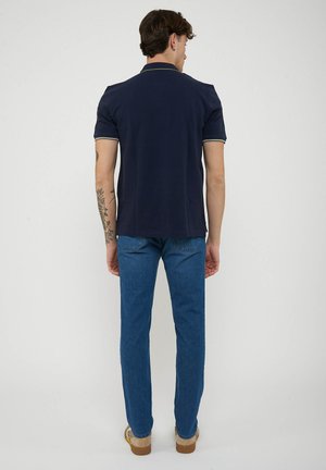 Uomo con le braccia tatuate in piedi di spalle che indossa una polo blu navy, jeans blu e scarpe beige su uno sfondo bianco semplice.