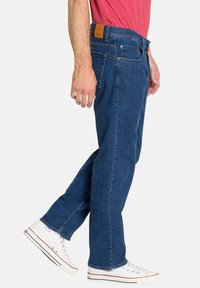 Jean en denim bleu à coupe droite, avec une étiquette en cuir à l'arrière de la taille, cinq poches et une fermeture éclair. Porté avec des baskets blanches.