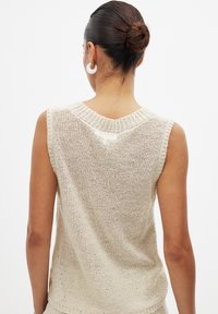 Haut en tricot sans manches de couleur beige avec un col côtelé, une texture en maille ouverte et un léger éclat. Design simple et sans ornements.
