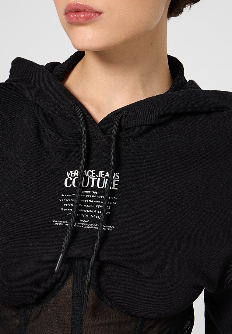 Sudadera negra con un gran bolsillo frontal, que presenta un texto de logo blanco y cordones. La tela parece suave, con un diseño en capas.