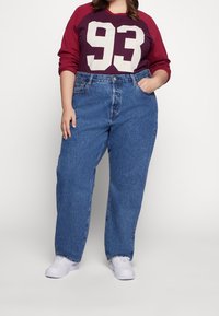 Camisa de manga larga color granate con estampado blanco "93", combinada con jeans de mezclilla azul de talle alto. La modelo lleva zapatillas blancas, de pie con una mano en la cadera.