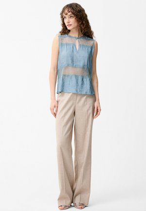 Femme aux cheveux bouclés portant un haut bleu sans manches avec des détails en dentelle et un pantalon beige à larges jambes, debout devant un fond blanc uni.