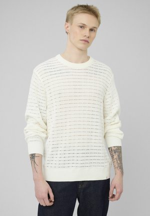 Jeune personne aux cheveux blonds courts portant un pull en maille blanche perforée et un jean foncé, montrant des tatouages sur les deux avant-bras.