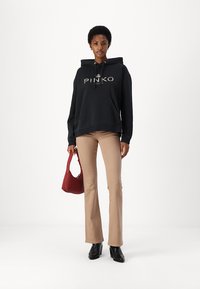 Zwarte hoodie met "PINKO" print in goud, beige flared broek, zwarte enkellaarsjes en een rode handtas. Eenvoudig ontwerp, casual stijl.