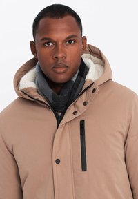 Giacca esterna beige con cappuccio removibile, caratterizzata da cerniere e bottoni neri, foderata in pile bianco, indossata sopra una sciarpa scura.