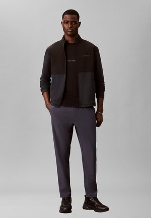 Ein erwachsener Mann steht und trägt eine schwarze Zip-Up-Jacke, ein schwarzes T-Shirt, dunkelgraue Hosen und schwarze Sneaker vor einem schlichten grauen Hintergrund.
