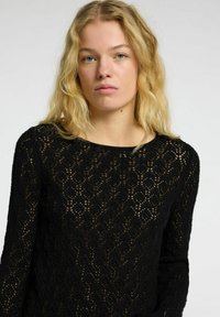 Maglione nero in maglia caratterizzato da un design in pizzo con motivi, scollatura rotonda e maniche lunghe. La consistenza è leggera e leggermente trasparente.
