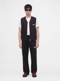 Homme portant un gilet noir sur un t-shirt blanc, pantalon noir avec logo blanc, lunettes de soleil sombres et chaussures foncées avec lacets rouges, debout devant un fond uni.