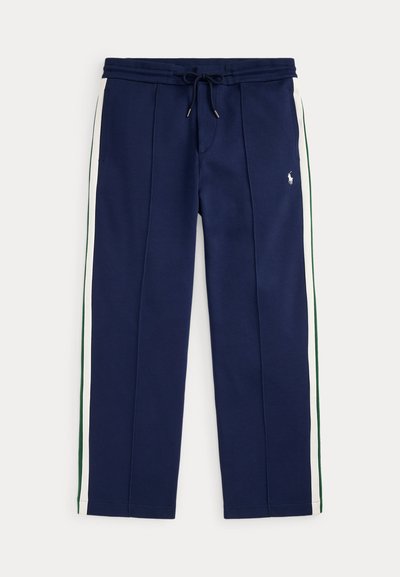 Pantalon jogger bleu marine avec des bandes latérales blanches et vertes, taille à cordon, poches avant, et petit logo blanc brodé sur la cuisse droite.