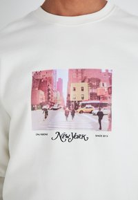 Sweat-shirt en coton blanc avec un graphisme d'une scène de rue vintage de New York, texte noir et encolure ronde.