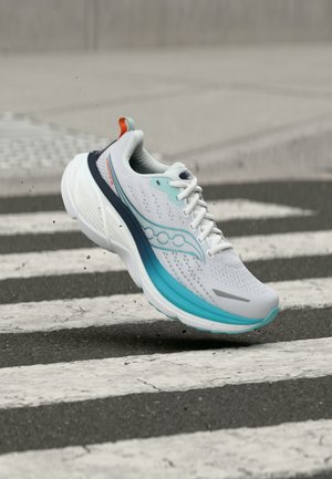 Valkoinen ja turkoosi juoksukenkä ilmassa mustavalkoisen suojatien yllä asfalttitiellä.