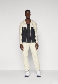 Treningsantrekk som inkluderer en svart og krem zip-up jakke, svart tank-top, kremfargede bukser og hvite joggesko med blå detaljer.