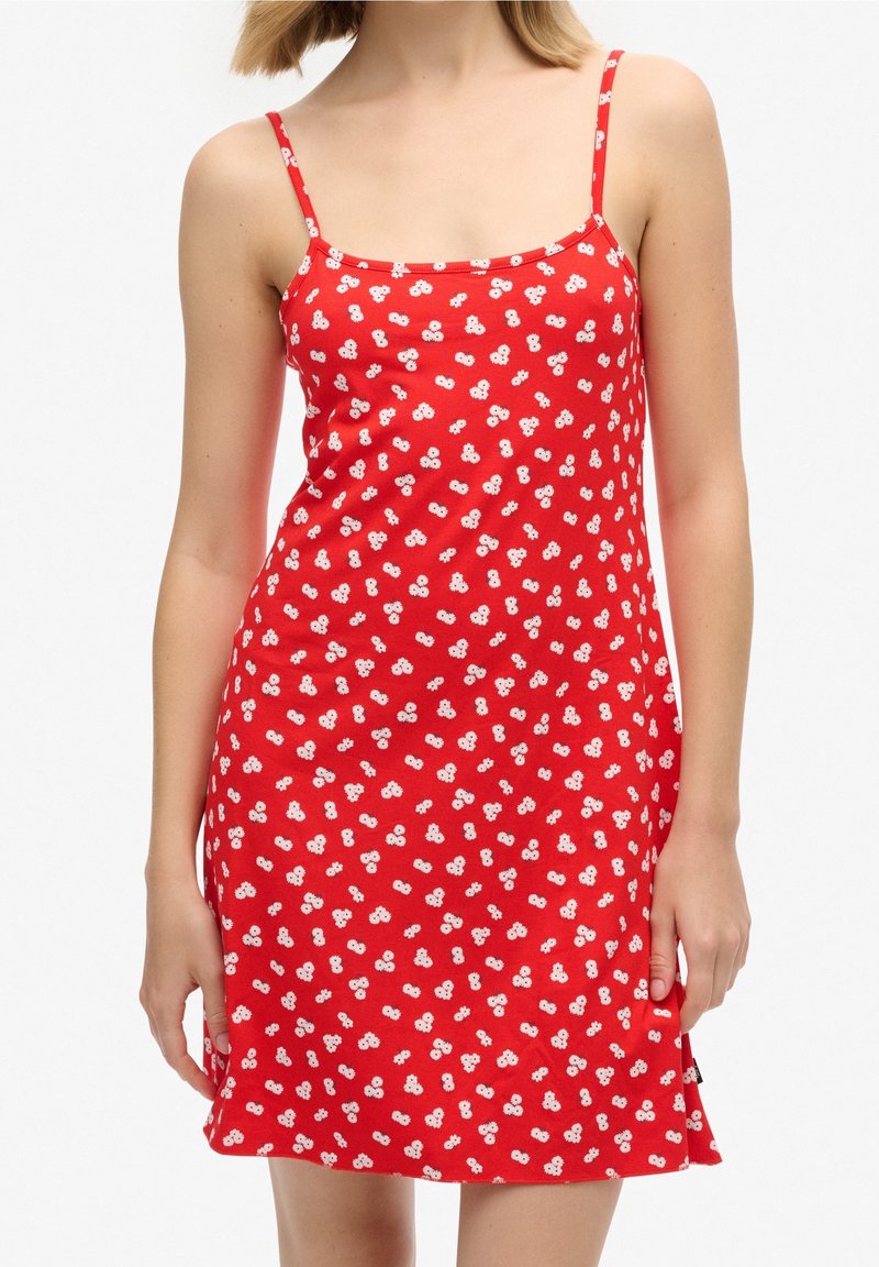 Femme portant une robe rouge sans manches avec de fines bretelles et un motif floral blanc, debout, les bras relâchés le long du corps.