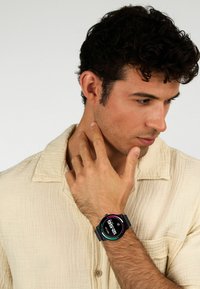 Orologio digitale nero con quadrante circolare, display colorato e cinturino testurizzato, indossato su un polso contro una camicia di colore chiaro.