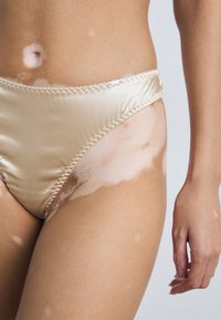 Bas de bikini en satin beige avec un design échancré, une taille élastique et des coutures délicates. Présente une peau avec des zones plus claires.