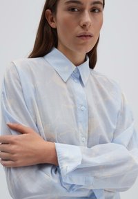 Camicia leggera azzurra trasparente con colletto, chiusura frontale con bottoni e motivo floreale. Maniche lunghe con polsini abbottonati e vestibilità ampia.