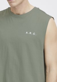 Ærmeløs olivengrøn T-shirt, lavet af blødt bomuld, med de hvide bogstaver "A.R.C." på venstre bryst. Glat tekstur, afslappet design.