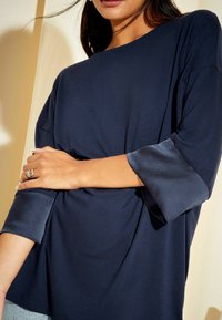 Mörkblå topp med avslappnad passform, med satinärmar i 3/4-längd. Tyget har en slät yta. Dekorativ ring på handen.