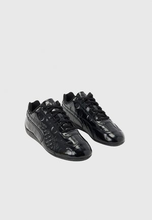 Par skinnende sorte sneakers med præget cirkulært mønster og sorte snørebånd på en ensfarvet lysegrå baggrund.