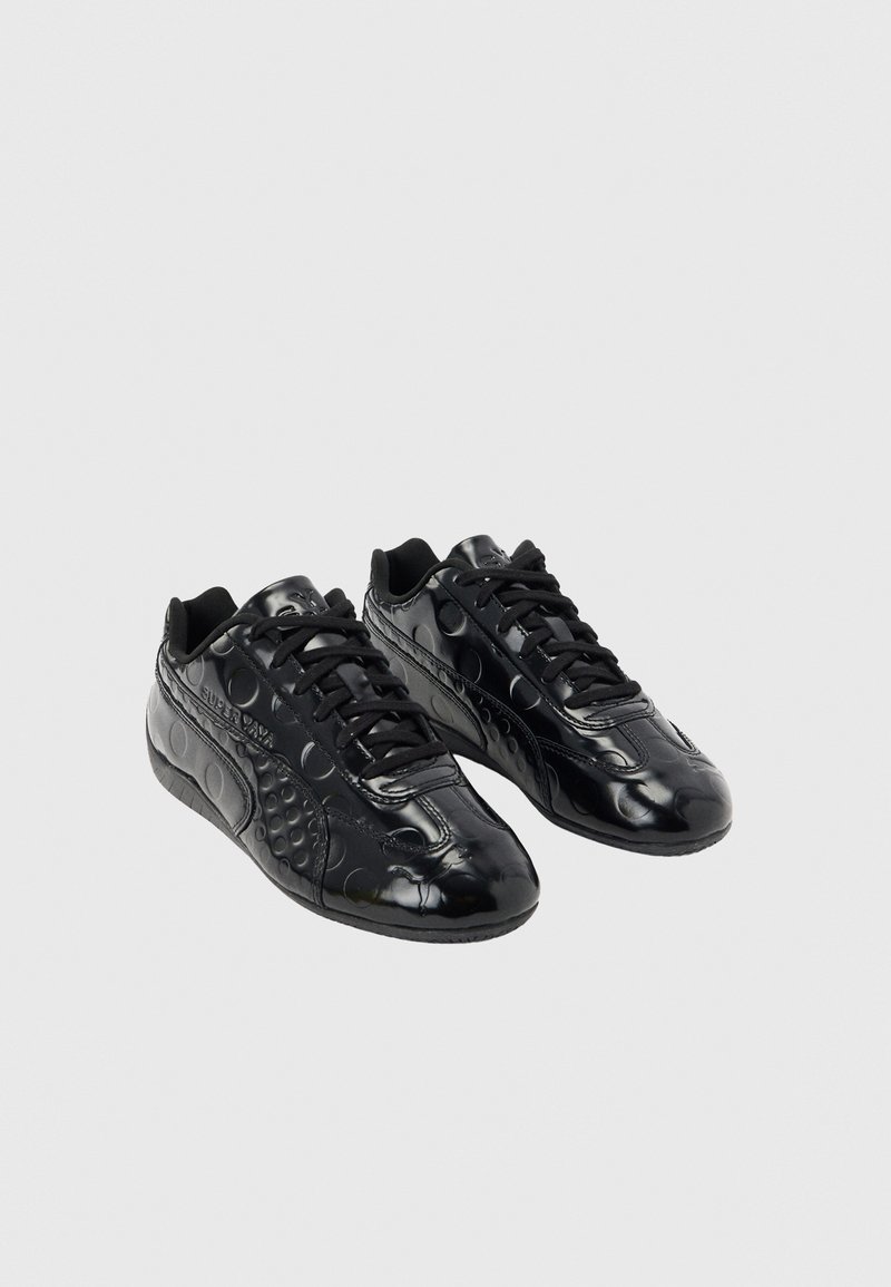 Paire de baskets noires brillantes avec motifs circulaires en relief et lacets noirs sur un fond gris clair uni.