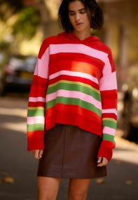 Hoodie à rayures rouges avec des accents roses, verts et blancs ; associé à une jupe en cuir marron. Texture douce ; coupe décontractée.