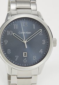 Sølv rustfritt stålklokke med en mørkeblå teksturert urskive, hvite time markører og en datovisning. Kjekk, glatt design med armbåndsrem.