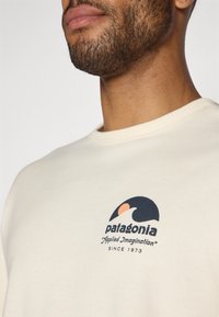 Kermanvärinen collegepaita pyöreällä pääntiellä ja resoroiduilla resoreilla hihoissa. Edessä tummansininen ja korallinpunainen graafinen logo, jossa lukee "patagonia" ja teksti "Applied Imagination" vuodesta 1973.