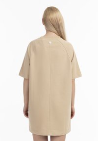 Beige oversized t-shirtklänning med korta ärmar, rund halsringning och knapppdetailj på baksidan; mjukt tyg med en slät textur.
