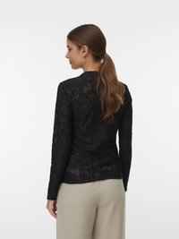 Hosszú barna hajú nő lófarokba kötve, fekete csipkés, hosszú ujjú blúzt és bézs nadrágot viselve, háttal áll egy sima fehér háttér előtt.