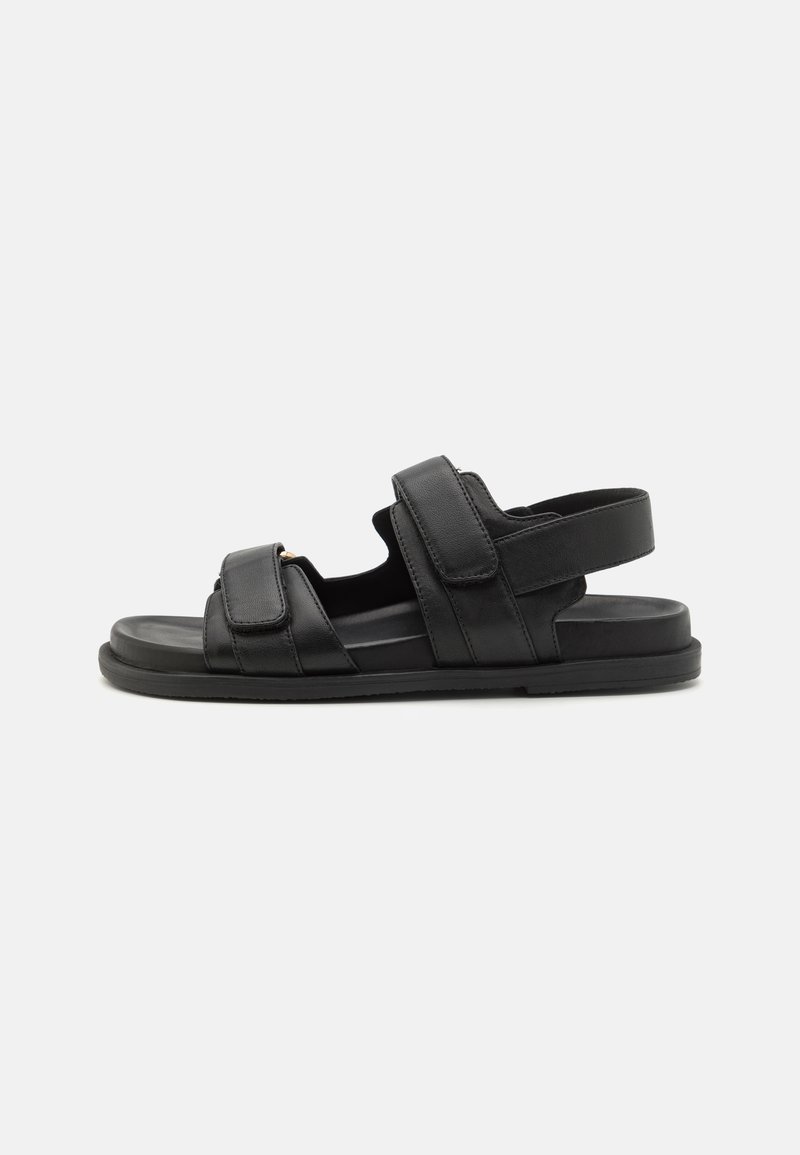 Mustat nahkasaappaat, joissa on säädettävät Velcro-hihnat, avonainen kärki ja teksturoitu pohja pidon parantamiseksi. Ominaisuudet: minimalistinen tyyli.