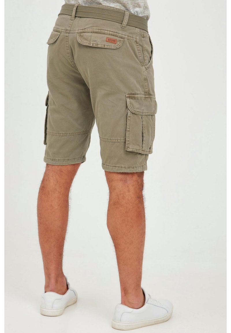Homme portant un short cargo beige avec plusieurs poches et des baskets blanches, vu de dos sur un fond uni.