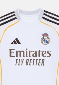 Maillot de sport blanc avec col gris, accent noir et jaune, arborant le logo du Real Madrid et "Emirates FLY BETTER" en noir.