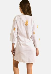Robe en coton blanche à manches trois-quarts, ornée d'une broderie jaune vive sur les épaules et le dos, avec des fentes sur les côtés pour faciliter les mouvements.