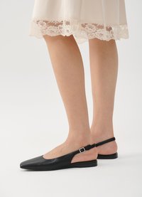 Chaussures plates noires à talons slingback avec un bout pointu, en cuir lisse, et un accent en boucle argentée. Portées avec une jupe crème bordée de dentelle.