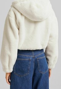 Fleecetröja i off-white med avslappnad passform, stor huva och elastiska ärmmuddar, tillsammans med högmidjade blåjeans.