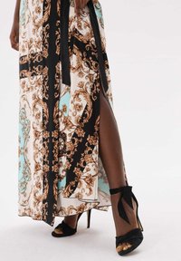 Robe maxi avec un motif floral beige et noir ornée sur un fond bleu clair, dotée d'une fente jusqu'à la cuisse. Sandales noires à talons avec des nœuds.