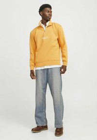 Felpa arancione con zip a metà e logo, abbinata a jeans azzurri chiari e scarpe marroni. Il modello è in piedi, rivolto leggermente a destra con le mani appoggiate.