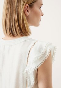 Blusa bianca con delicati dettagli in pizzo sui maniche e sulle spalle. Tessuto leggero e strutturato con dettagli a taglio orizzontale lungo le cuciture.
