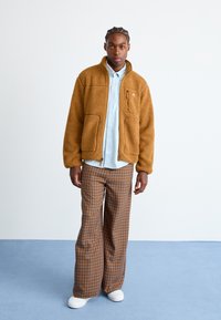Brown fleece jacket with zip closure and two front pockets, paired with wide plaid trousers in brown and orange, and white shoes.