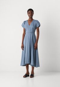 Lichtblauwe jurk met V-hals en korte pofmouwen, cinched taille, geplooide rok en asymmetrische zoom. Draag met zwarte instapschoenen.