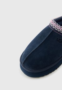 Σατέν παντόφλα σε ναυτικό μπλε χρώμα με ροζ κεντήματα κατά μήκος και ραμμένη κεντρική ραφή, με παχιά λαστιχένια σόλα και ανάγλυφο λογότυπο UGG.